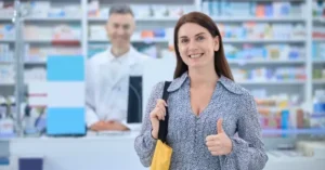 A smiling woman in a pharmacy with a thumb-up motion and a happy pharmacist standing behind a counter on her right.